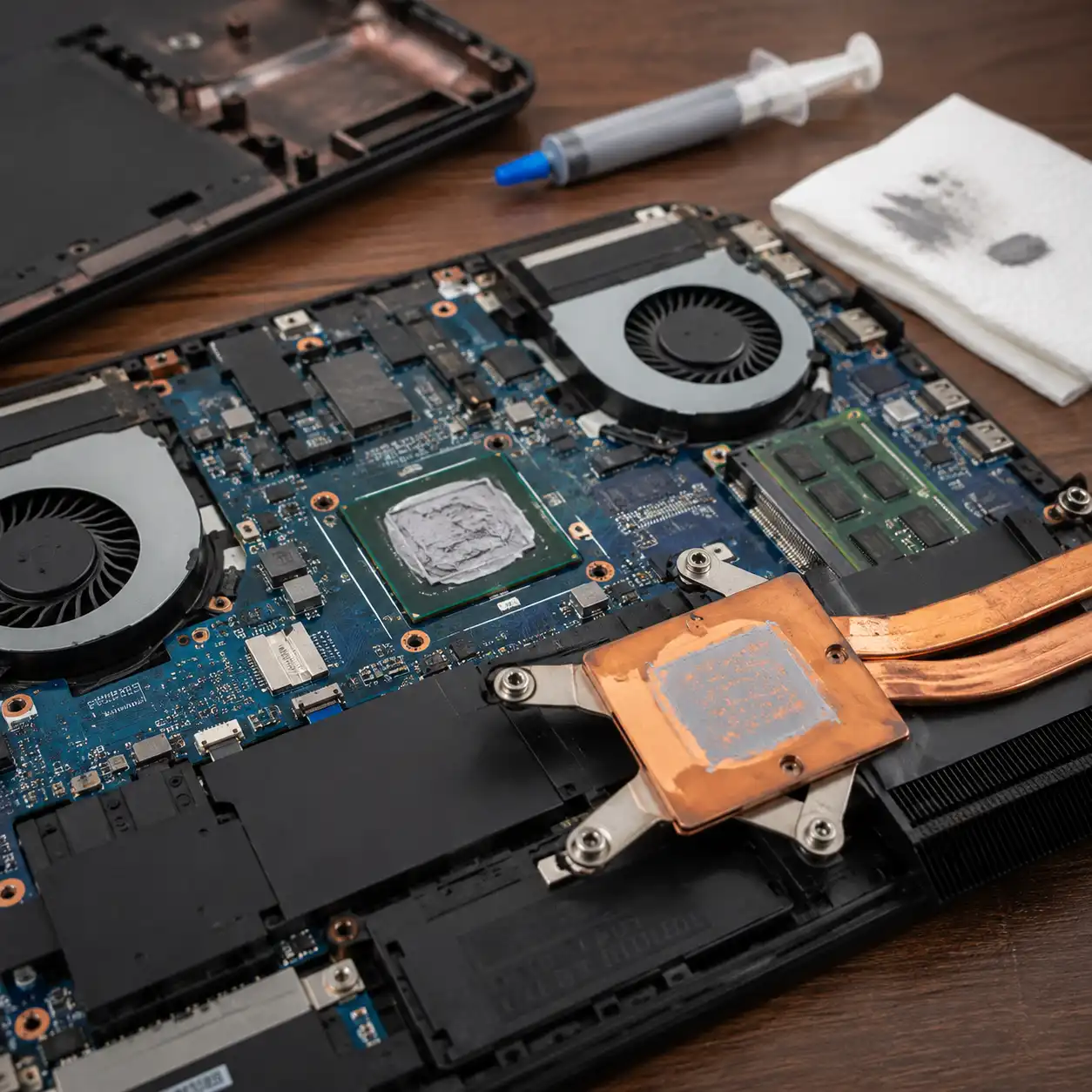 Laptop thermal paste replacement showing an open laptop, exposed processor, cooling fans, heatsink, and thermal paste syringe.